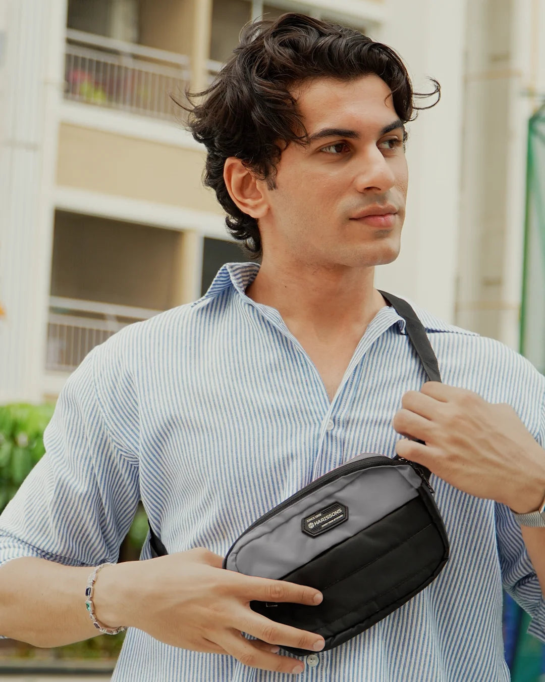 A man wearing a light blue and white striped button-down shirt models a black nylon crossbody bag with a silver zipper and a small black brand patch on the front, set against a blurred outdoor background.