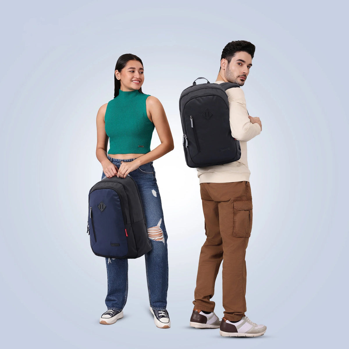 Two models wearing backpacks in a studio setting. The woman on the left holds a navy blue backpack, and the man on the right wears a black backpack.