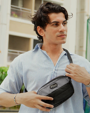 A man wearing a light blue and white striped button-down shirt models a black nylon crossbody bag with a silver zipper and a small black brand patch on the front, set against a blurred outdoor background.